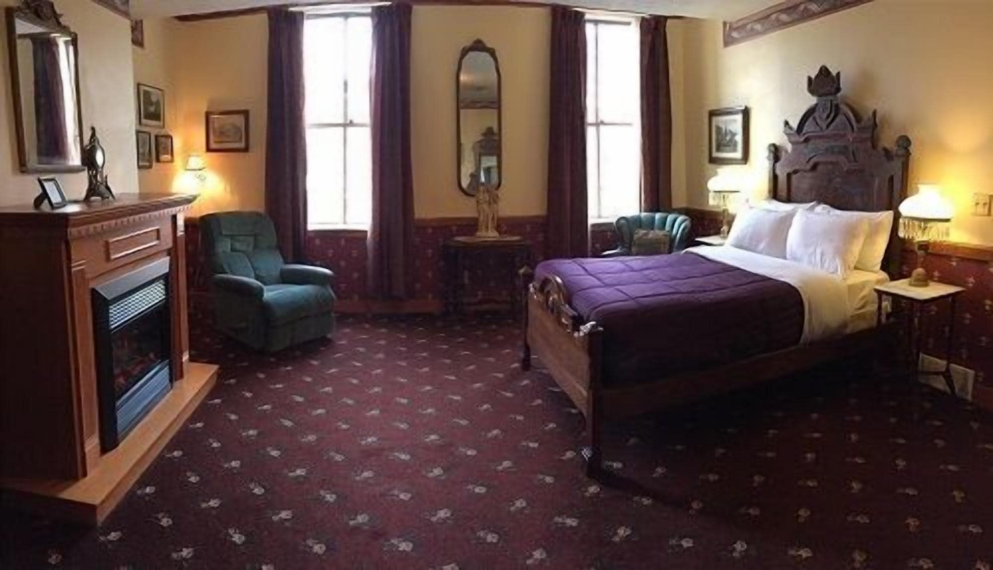 A hotel room with a large bed, a chair, a small table, a fireplace, and a mirror on the wall.