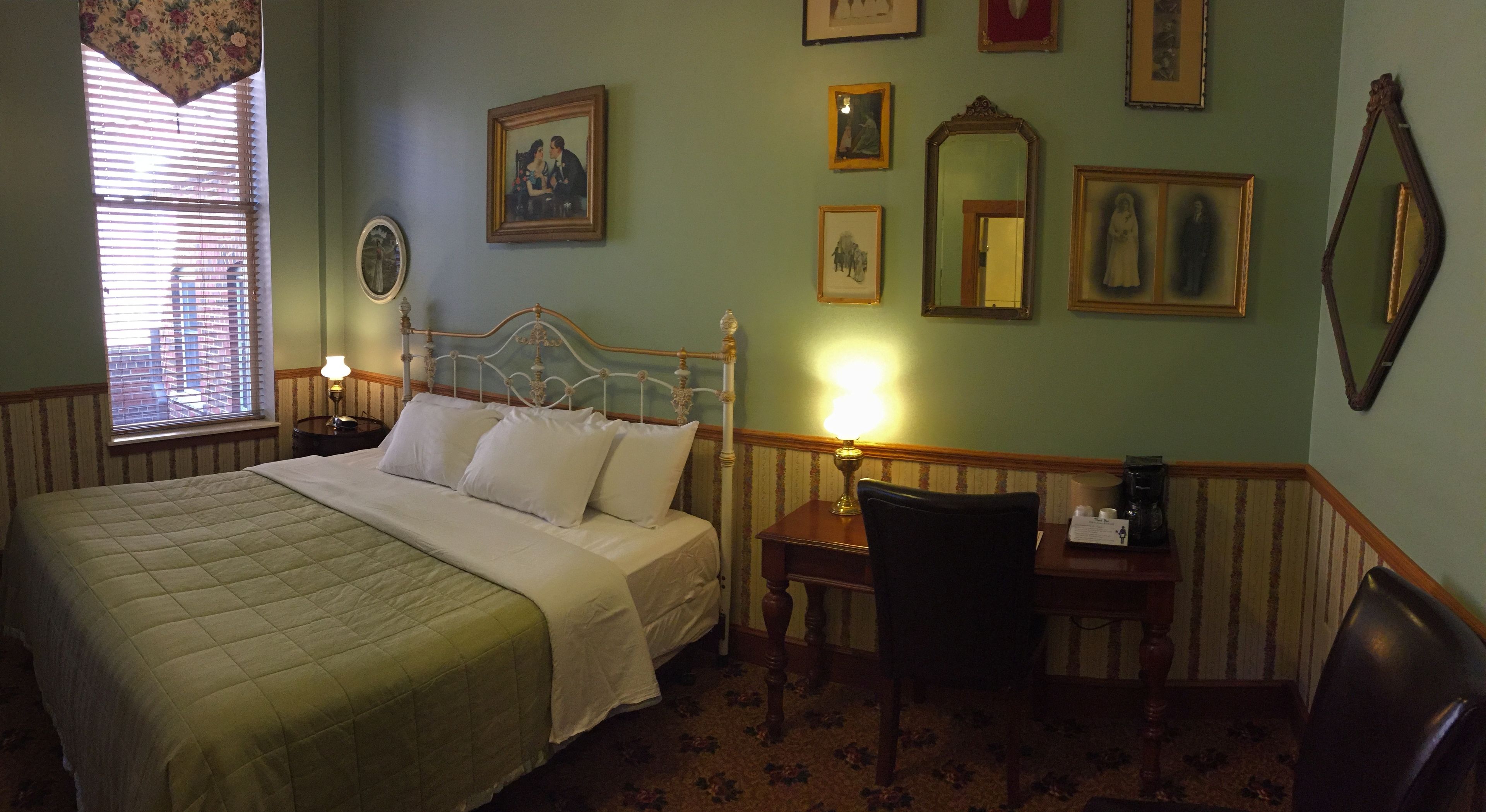 Habitación de hotel con cama, escritorio y silla. En la pared hay cuadros enmarcados y una ventana con persianas.