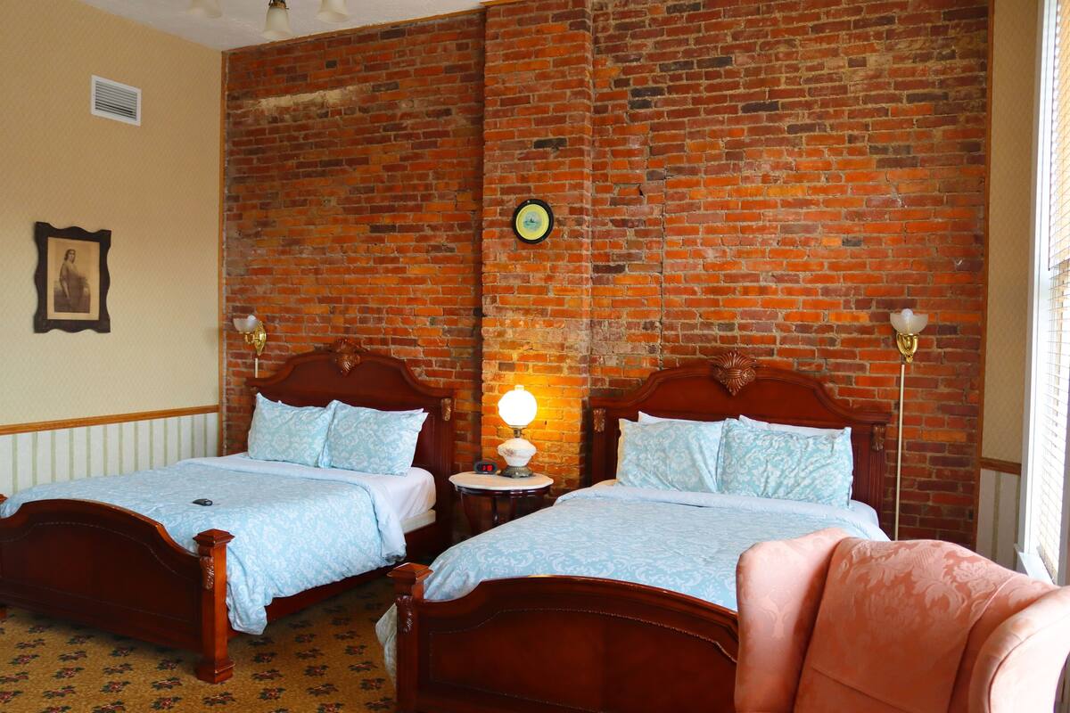 Ein Zimmer mit zwei Holzbett, einer Ziegelwand, einem kleinen runden Tisch mit einer Lampe und einem gerahmten Bild an der Wand.