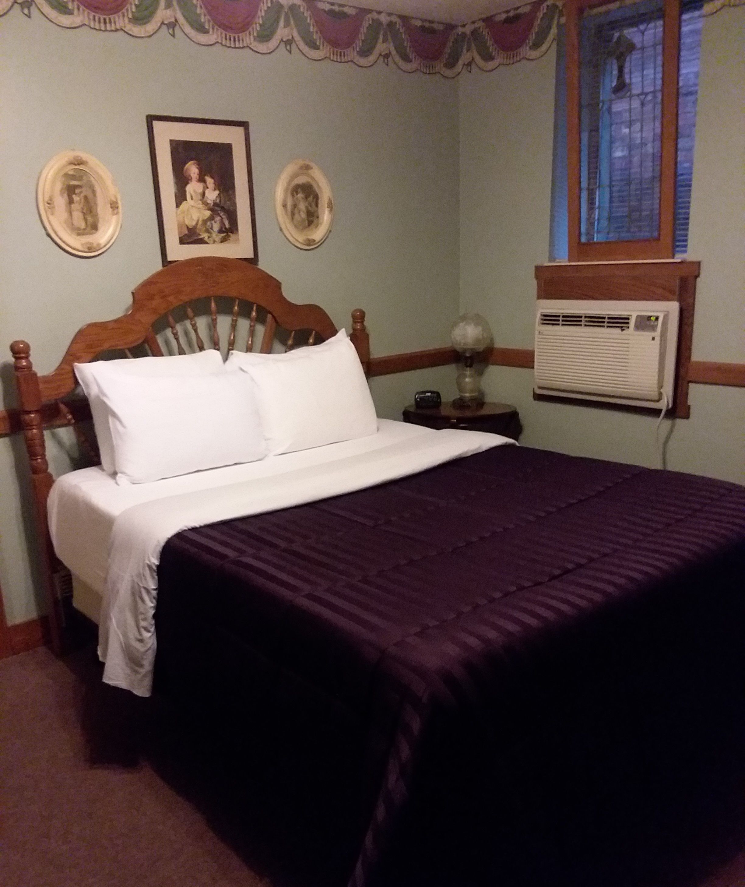 Un dormitorio con una cama de madera, ropa de cama blanca, una colcha morada, dos cuadros en la pared y una ventana con persianas.
