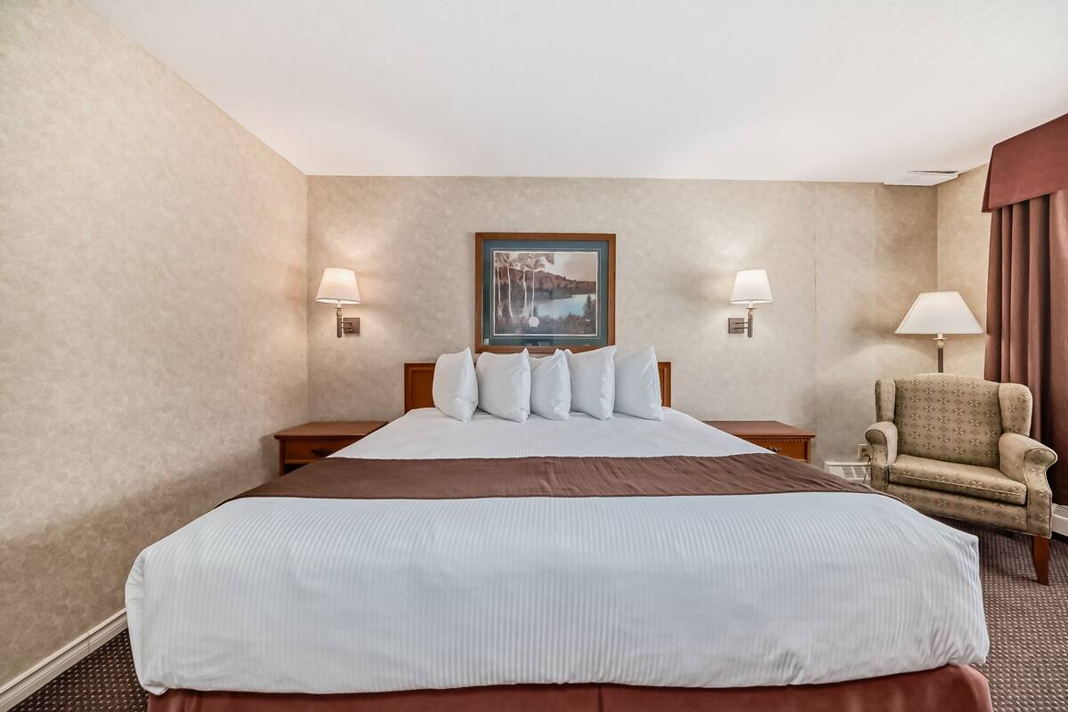 Une chambre d’hôtel avec un grand lit, deux lampes de chevet, un tableau encadré au mur et un fauteuil à motifs.