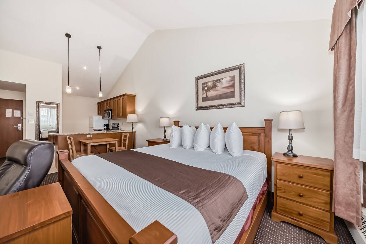 Une chambre d’hôtel avec un grand lit, une tête de lit en bois, des tables de chevet, un bureau et une kitchenette.