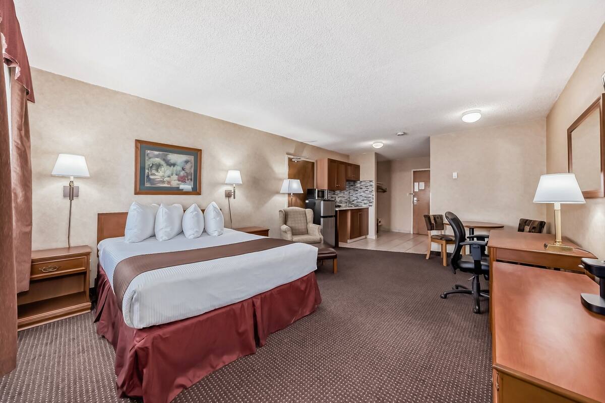 Une chambre d’hôtel avec un grand lit, un bureau, une chaise, un coin cuisine et une salle de bain visible à travers une porte ouverte.