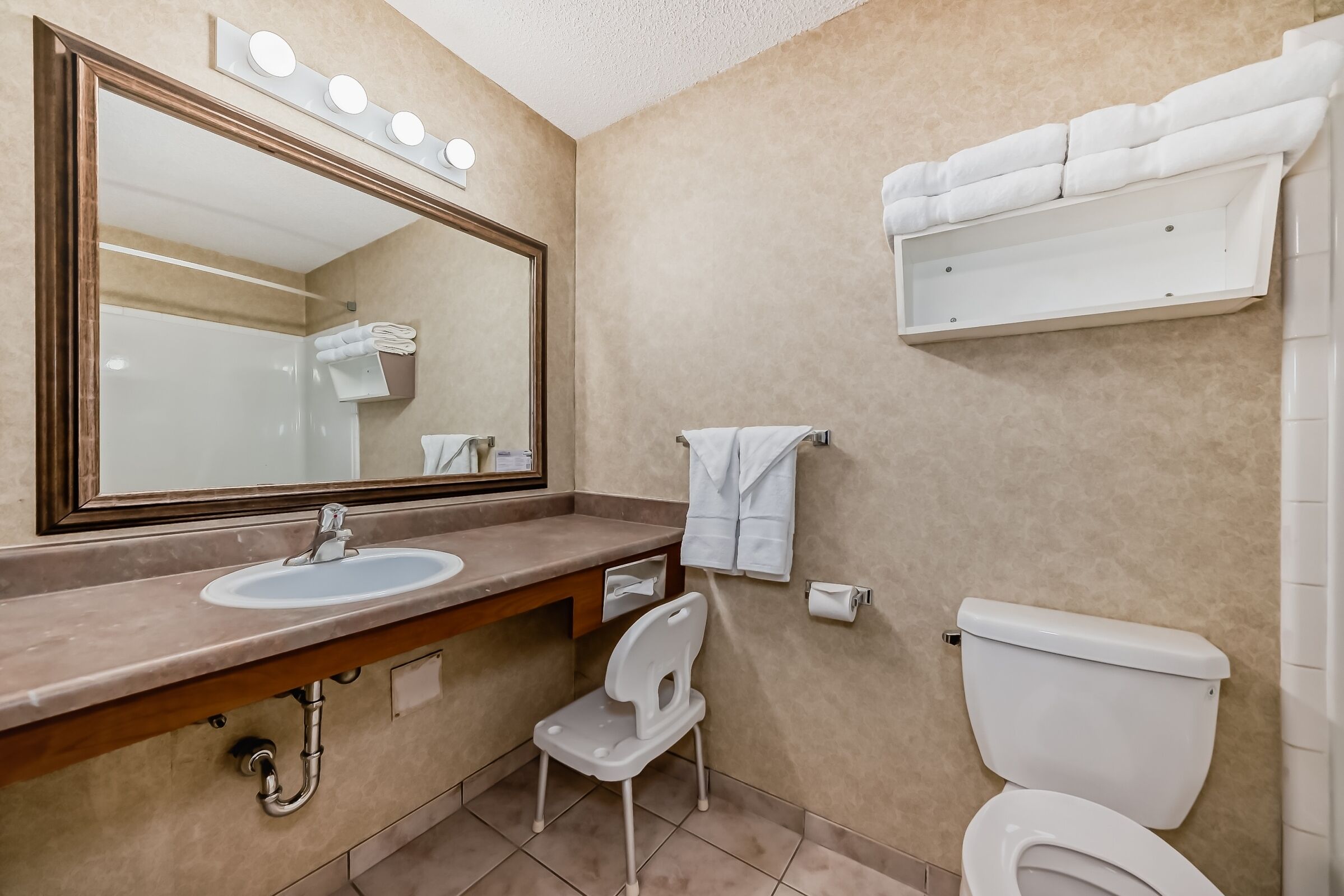 Ein Badezimmer mit Waschbecken, Spiegel, Handtuchhalter, Toilette und einem kleinen weißen Stuhl.