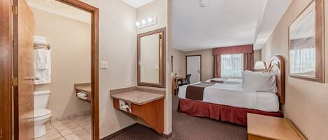 A hotel room with a bathroom, a bed with a red skirt, a vanity with a mirror, and a bedside table.