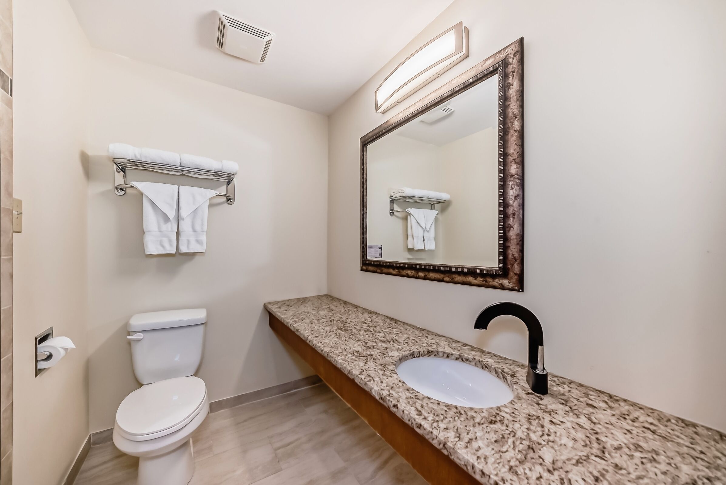 Un baño con inodoro, lavamanos con encimera de granito, un espejo grande con marco de madera y un perchero con dos toallas.