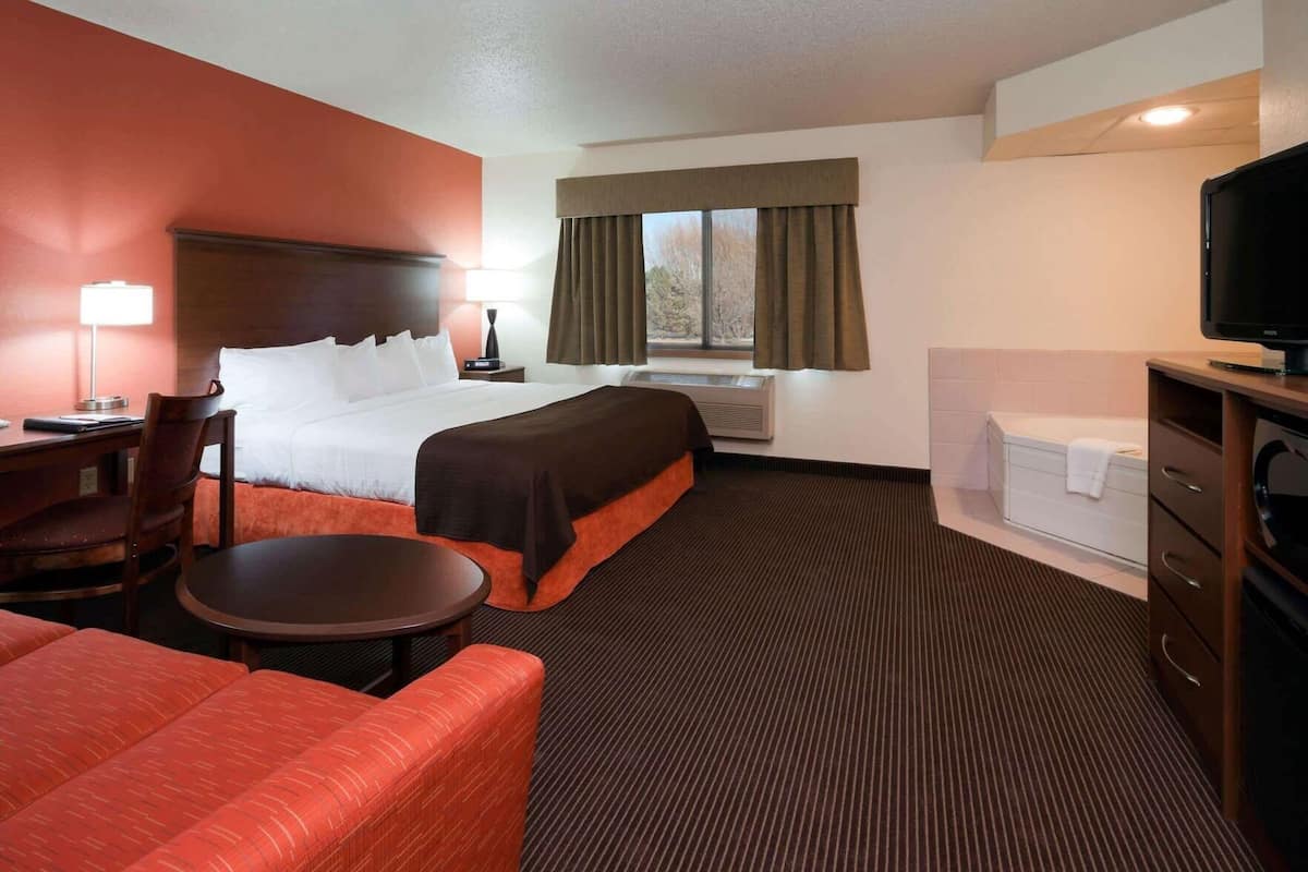 A hotel room with a bed, a desk, a chair, a small table, a TV, and a window with curtains.
