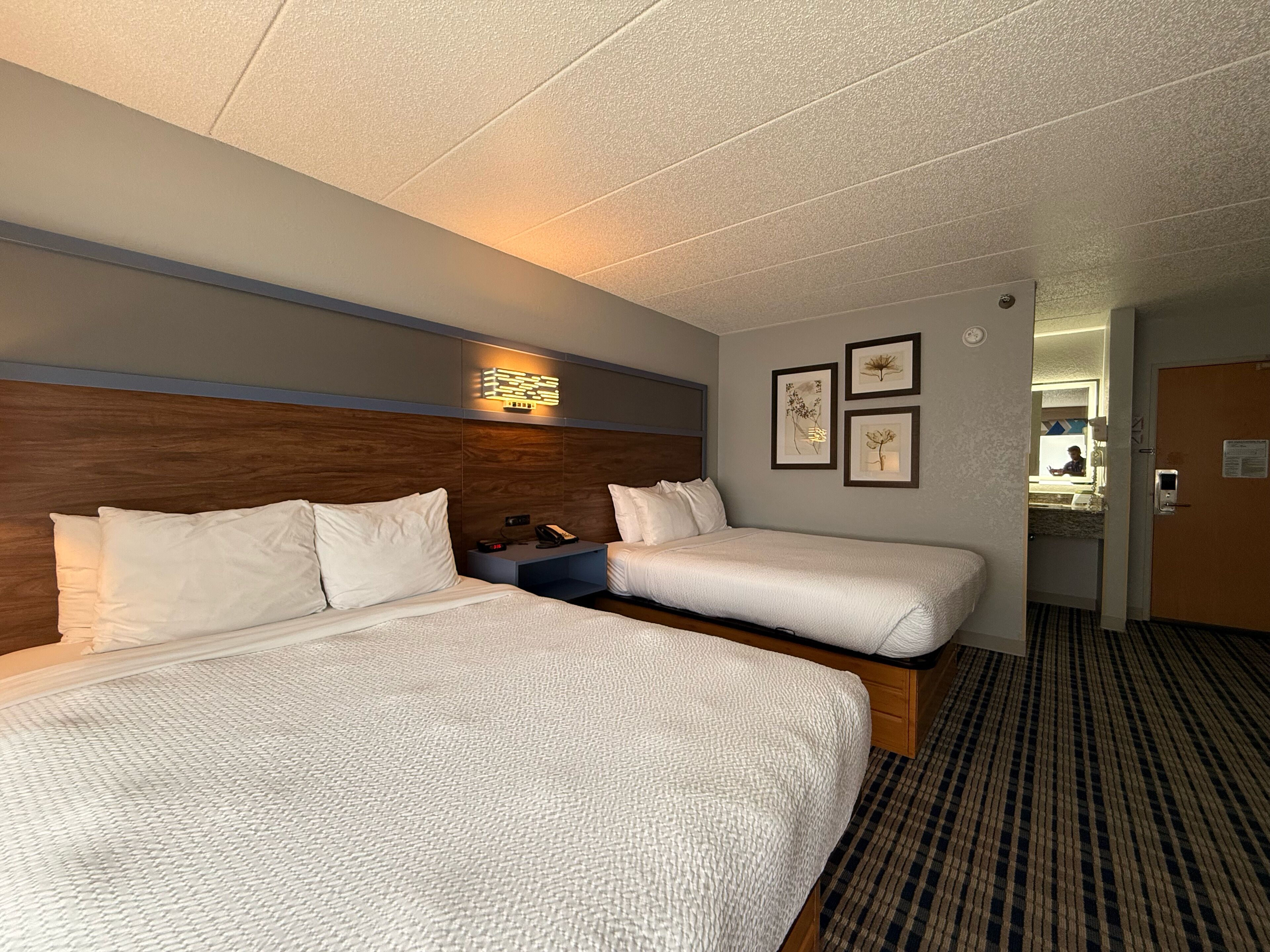A hotel room with two beds, a wooden headboard, a bedside table with a phone, and framed pictures on the wall.