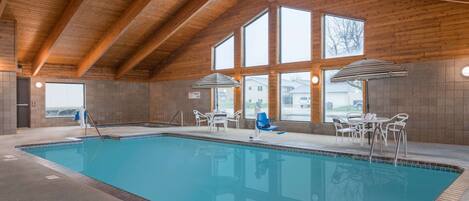 Una piscina interior con vigas de madera, ventanas amplias y sillas blancas alrededor de la piscina.
