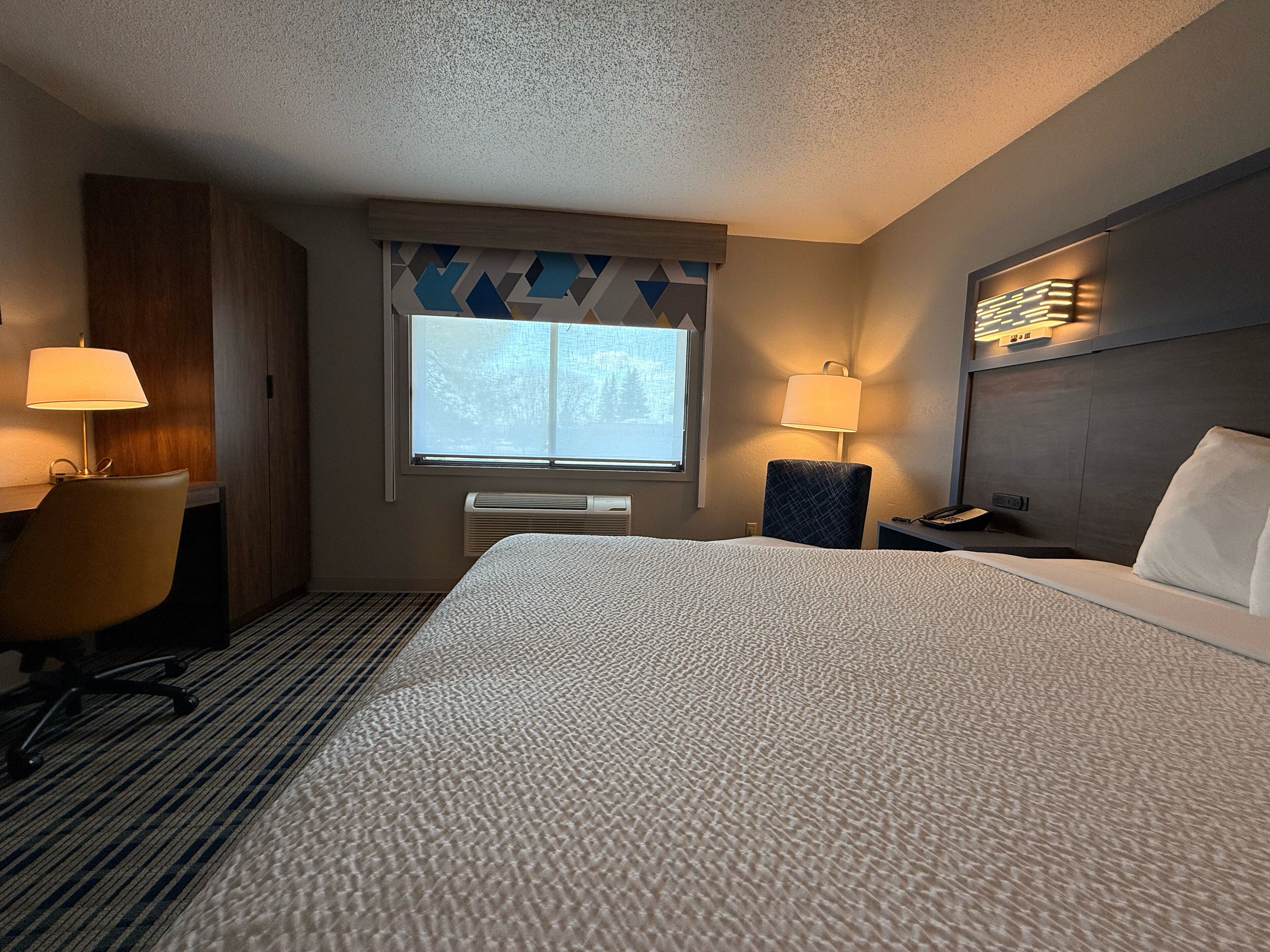 Habitación de hotel con una cama grande, dos mesitas de noche, un escritorio con una silla, una ventana con vistas y una lámpara de pared.