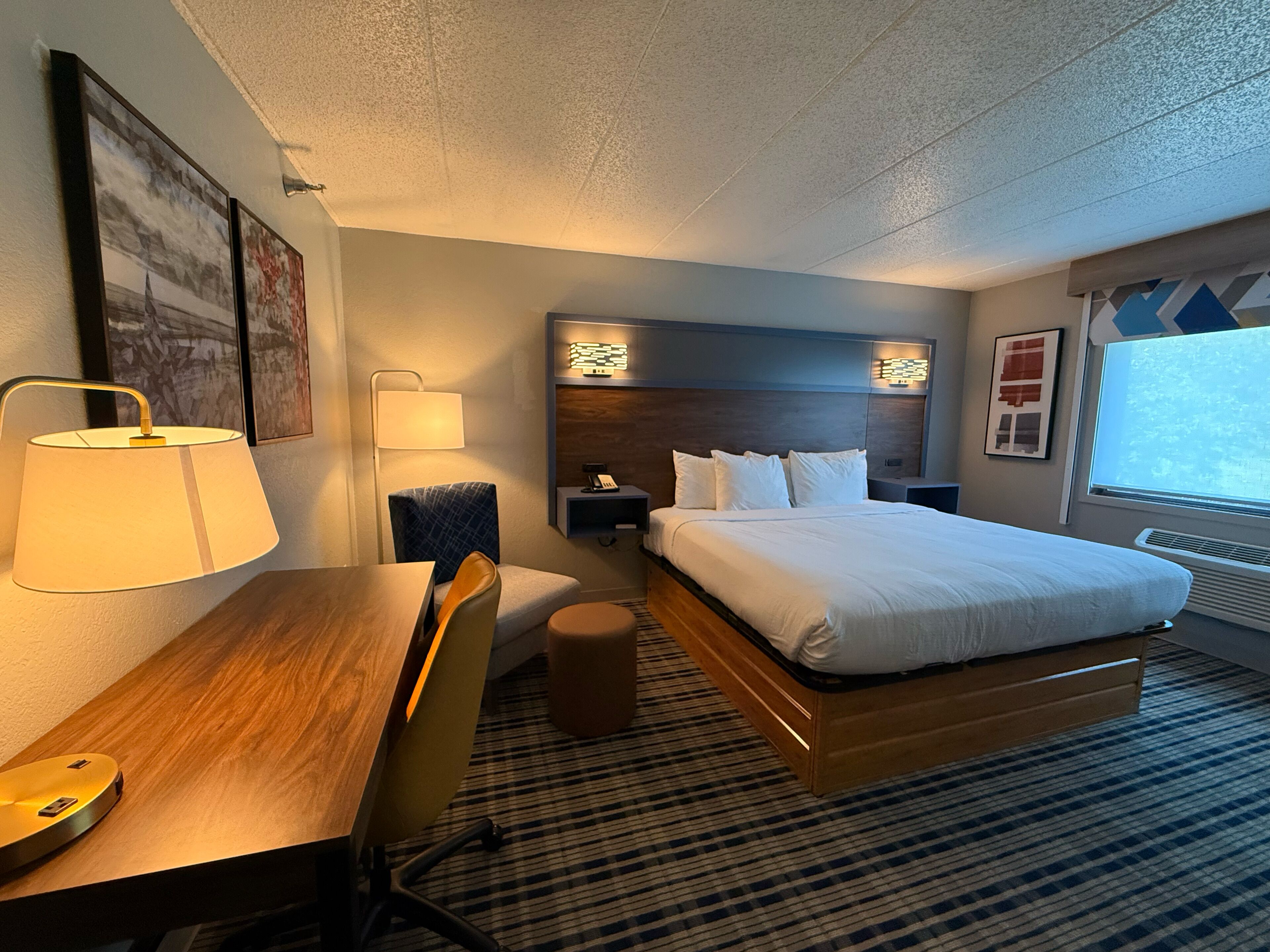 Una camera d'albergo con un letto, un comodino, una lampada, una sedia e una scrivania con un computer.