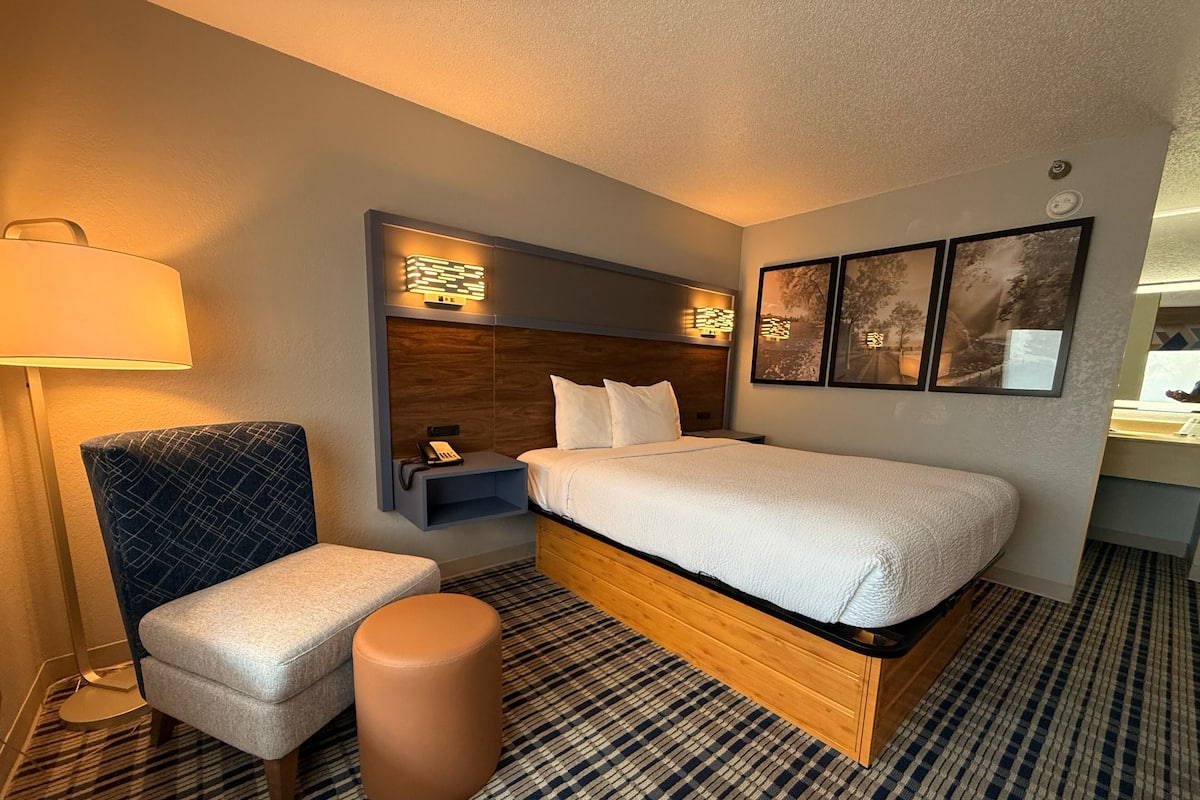 A hotel room with a bed, a chair, a small ottoman, a nightstand with a phone, and wall-mounted lights.
