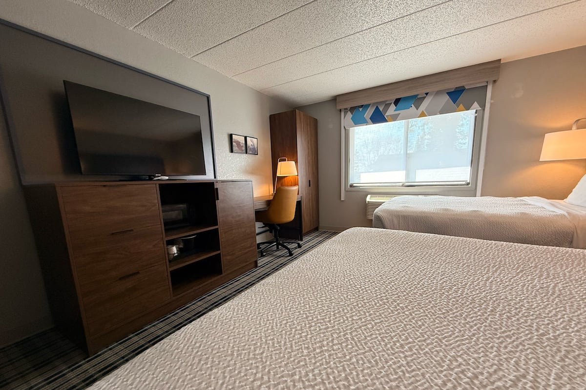 A hotel room with two beds, a TV, a desk, and a window with a view.
