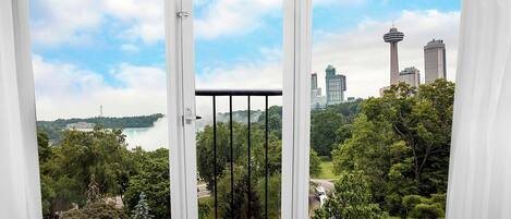 A view through a window with a white frame and black bars, overlooking a cityscape with tall buildings, including a tower with a circular observation deck, and a body of water with a distant building.