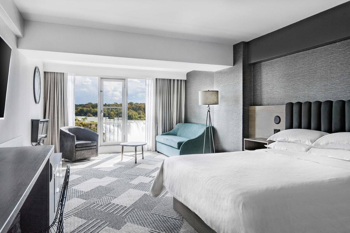 A modern hotel room with a large bed, a desk, a chair, a floor lamp, and a view of the outdoors.