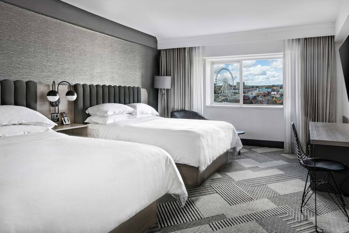 A hotel room with two beds, a desk, a chair, and a large window with a city view.