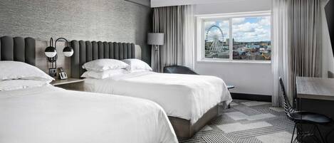 A hotel room with two beds, a desk, a chair, and a large window with a city view.