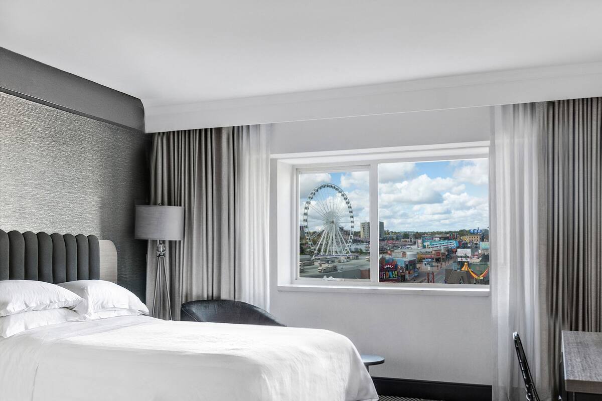 A hotel room with a bed, a chair, a desk, a lamp, and a window with a view of a Ferris wheel and buildings.