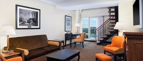 A hotel room with a living area featuring a sofa, armchairs, a coffee table, and a desk. There is a staircase leading to a balcony with a view.