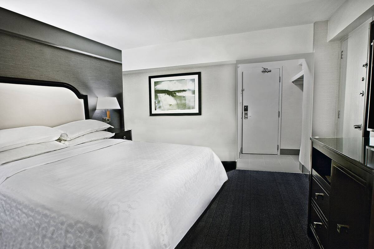 A hotel room with a large bed, a bedside table with a lamp, a framed picture on the wall, a wardrobe, and a mirror.