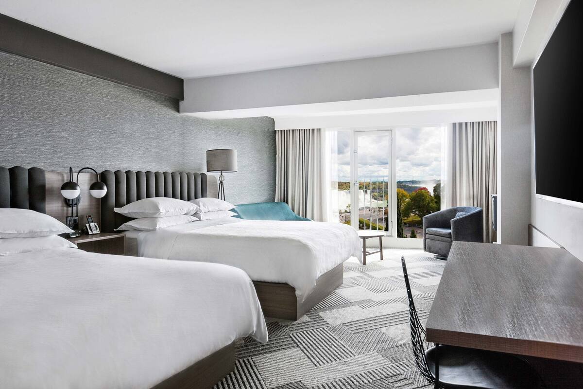 A modern hotel room with two beds, a dining table, and a balcony with a view.
