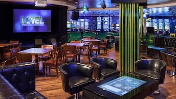 A sports bar and entertainment area with a large screen, gaming machines, leather seating, and a bar.