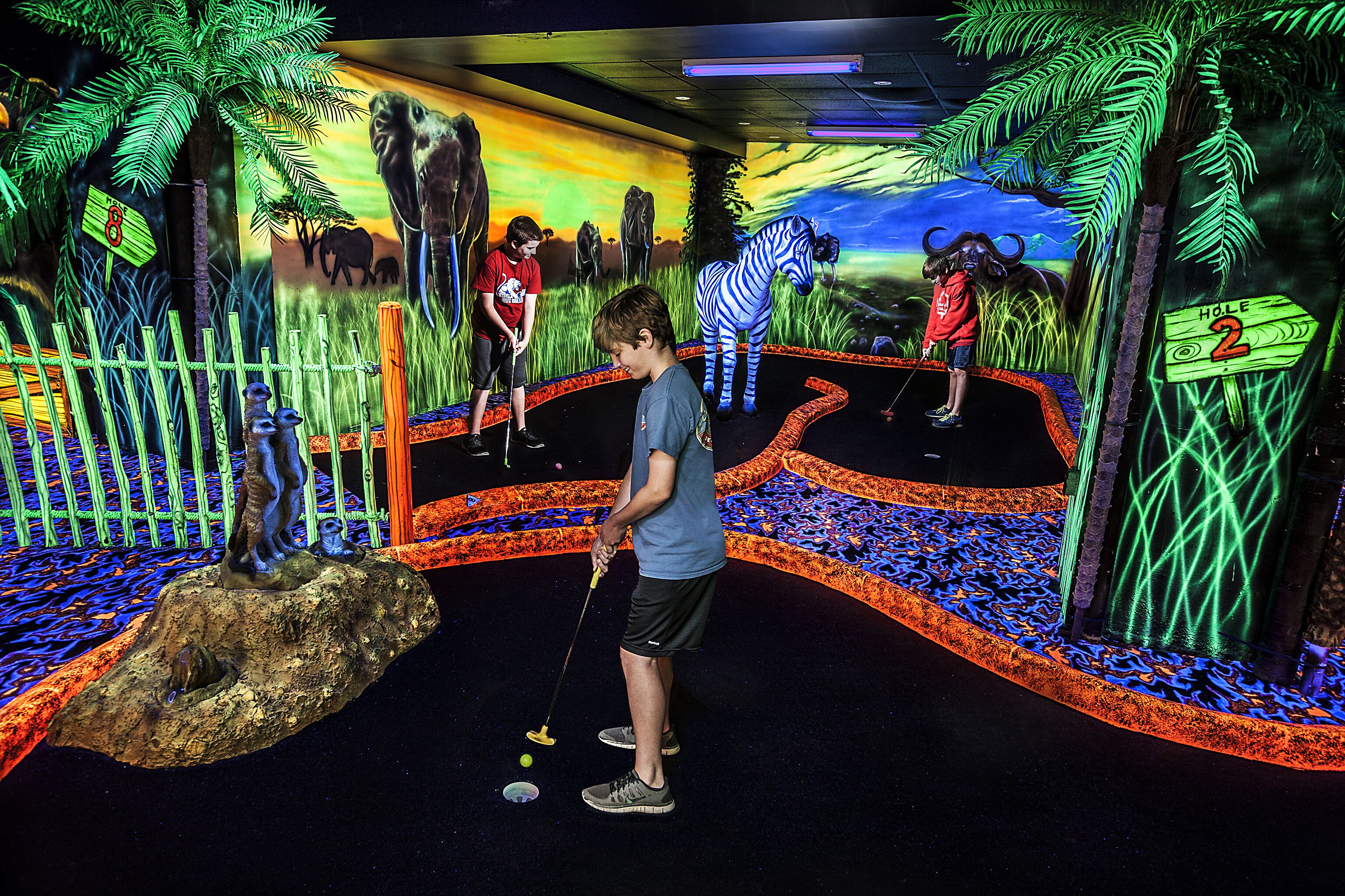 Un enfant joue au mini-golf dans une pièce décorée sur le thème de la jungle, avec une grande fresque représentant des animaux et des arbres.