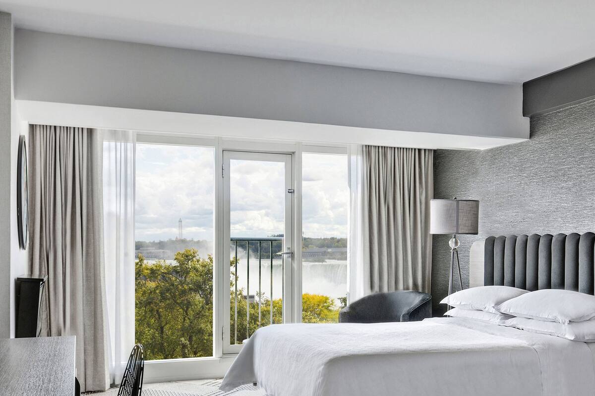A modern hotel room with a large window, a bed, a chair, and a lamp.