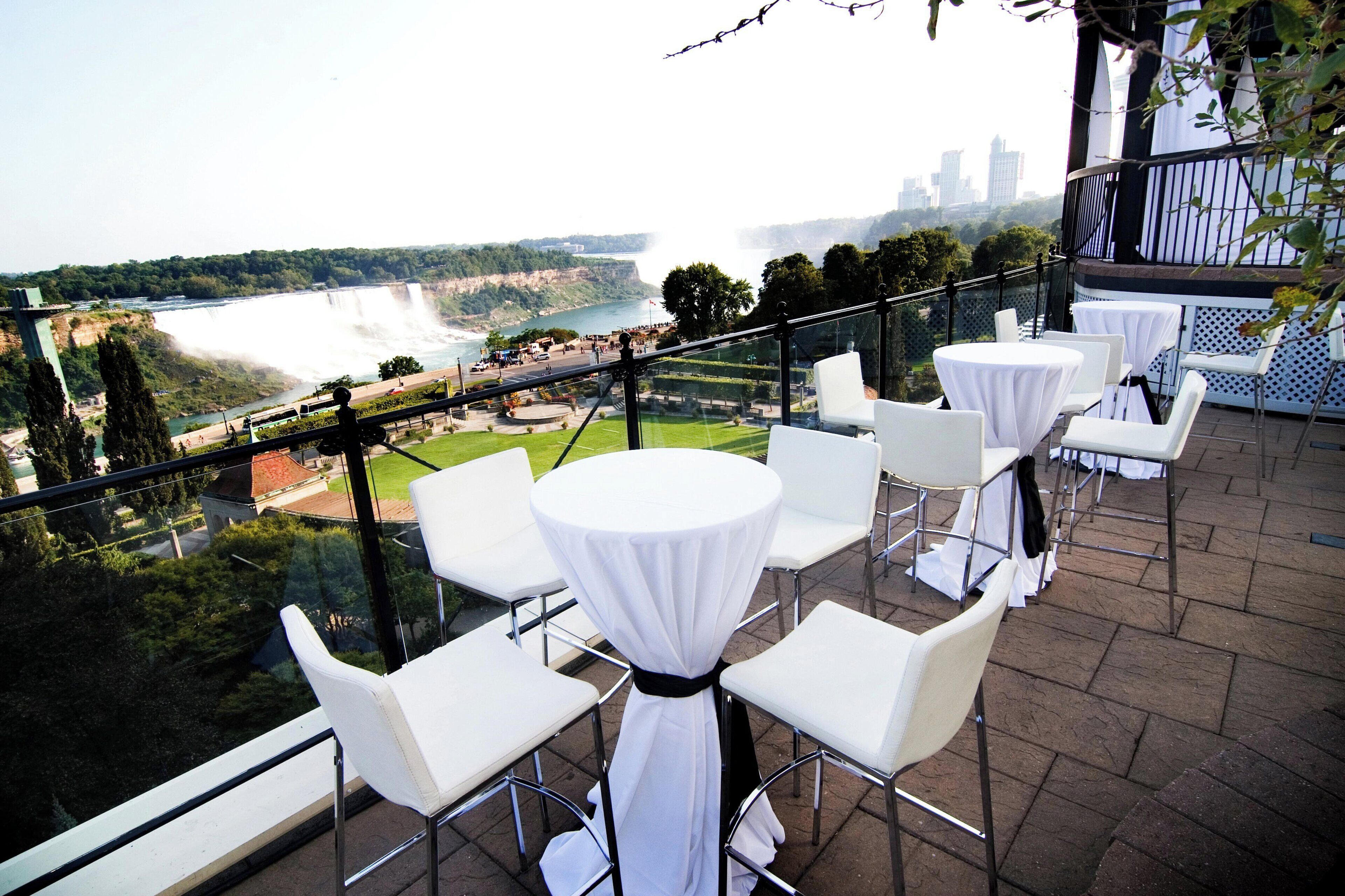 A terrace with white tables and chairs, overlooking a river and city skyline.