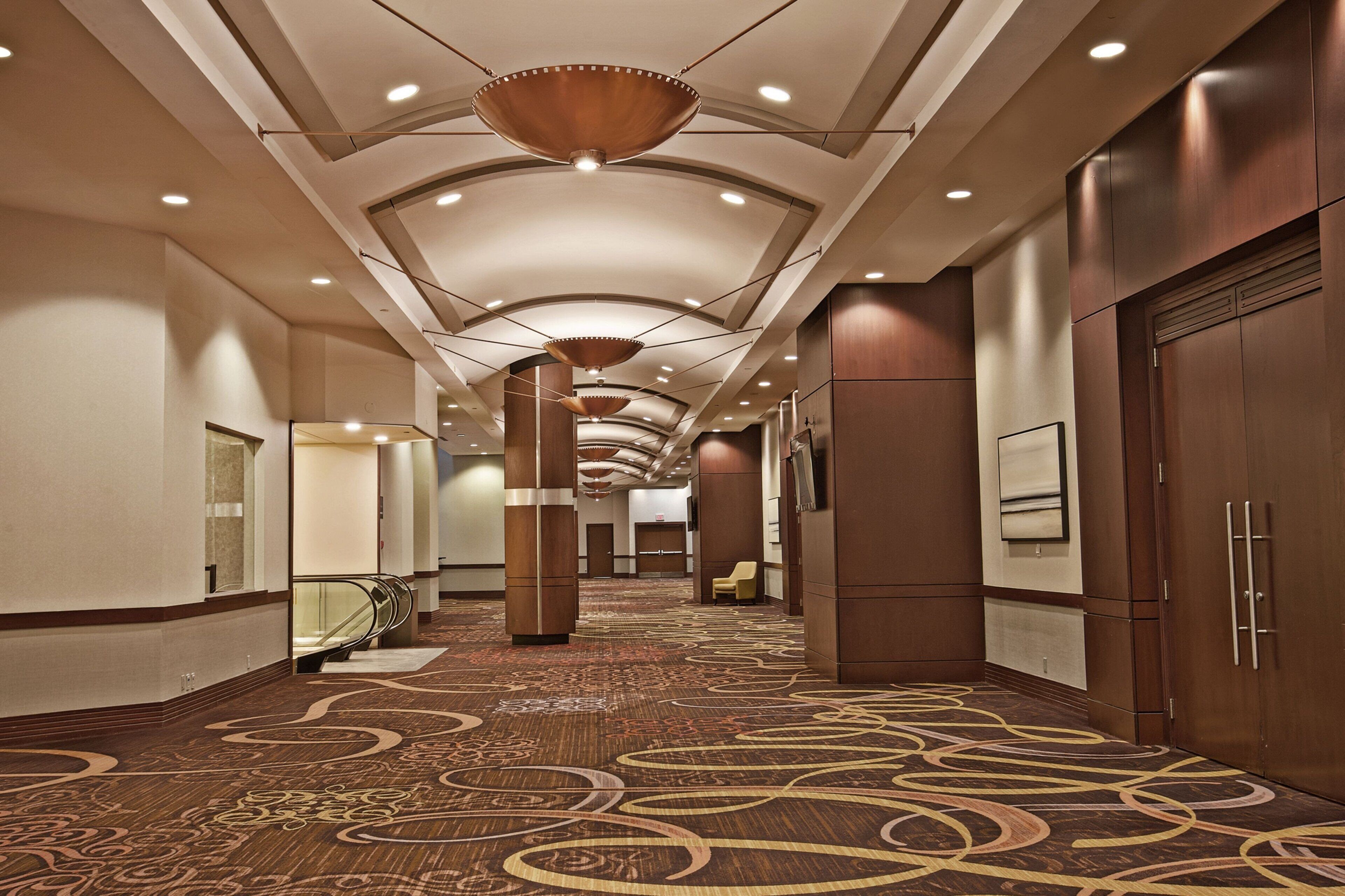 A hotel corridor with a patterned carpet, wooden paneling, and a modern ceiling design.