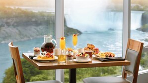 A breakfast table with a variety of dishes, drinks, and a pitcher, set against a backdrop of a scenic view of a waterfall.