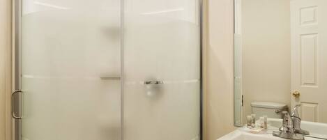 A glass-enclosed shower with a handheld showerhead, a white sink with a mirror, and toiletries.