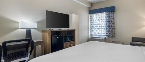 Hotellihuone, jossa on sänky, työpöytä, jossa on litteänäyttöinen televisio, mikroaaltouuni, jääkaappi, tuoli ja ikkuna, jossa on kuviolliset verhot.