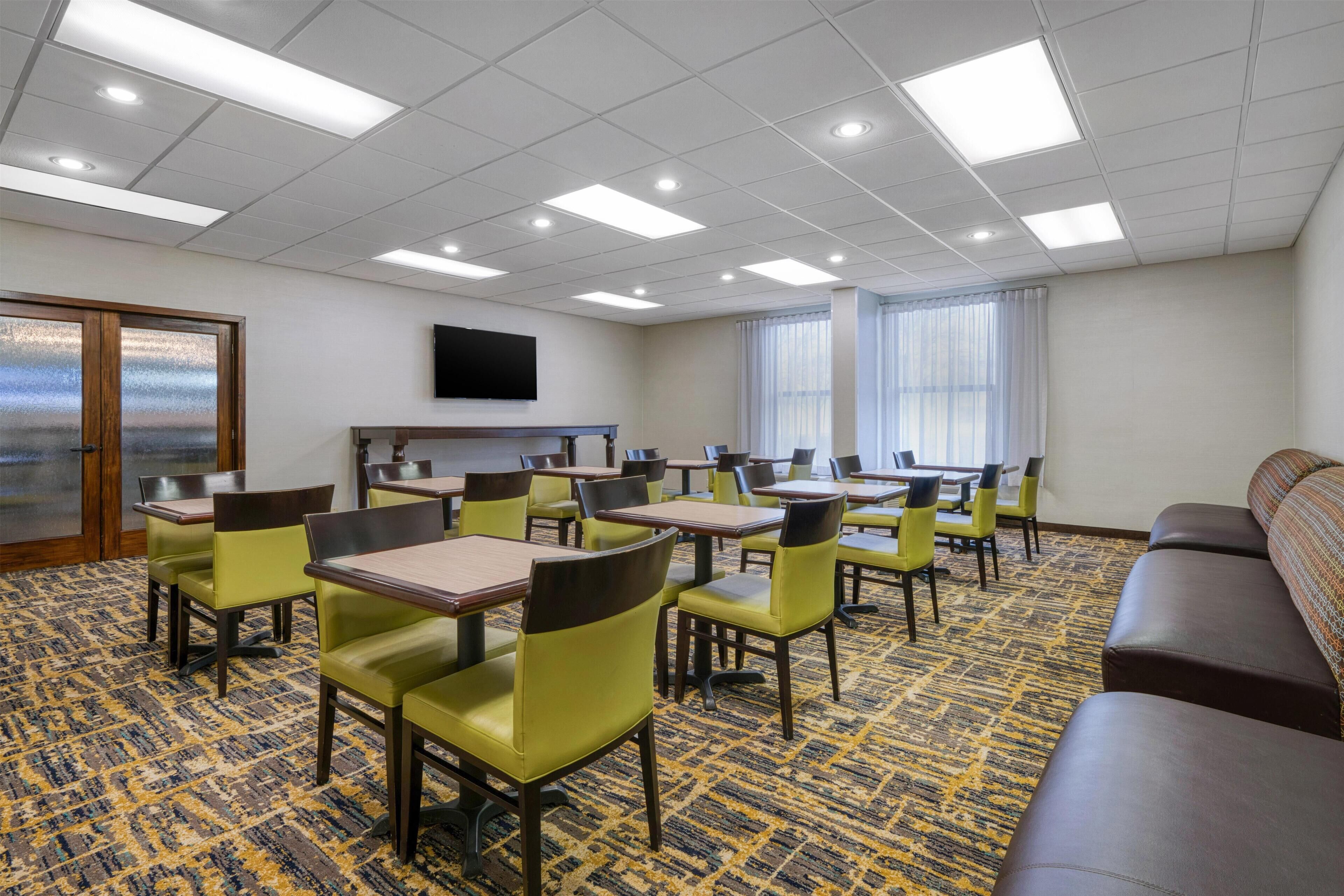 Habitación de hotel con piso alfombrado, varias mesas y sillas, un sofá, un televisor montado en la pared y una ventana con cortinas.