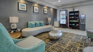 A modern lobby with a curved sofa, a round ottoman, a patterned rug, a refrigerator, a vending machine, and framed pictures on the wall. - Best Western Fishers/Indianapolis Area (Fishers)