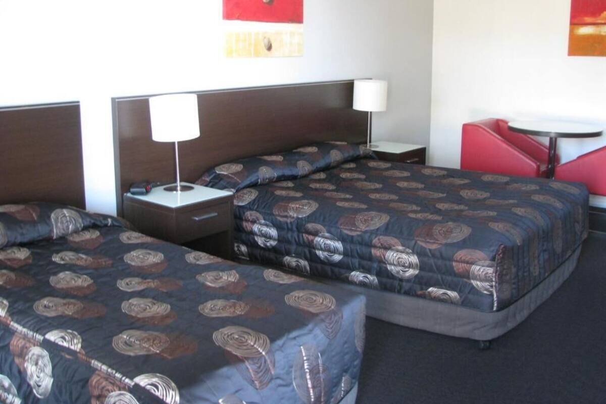 Quarto de hotel com duas camas, uma mesinha de cabeceira, uma luminária e uma cadeira vermelha.