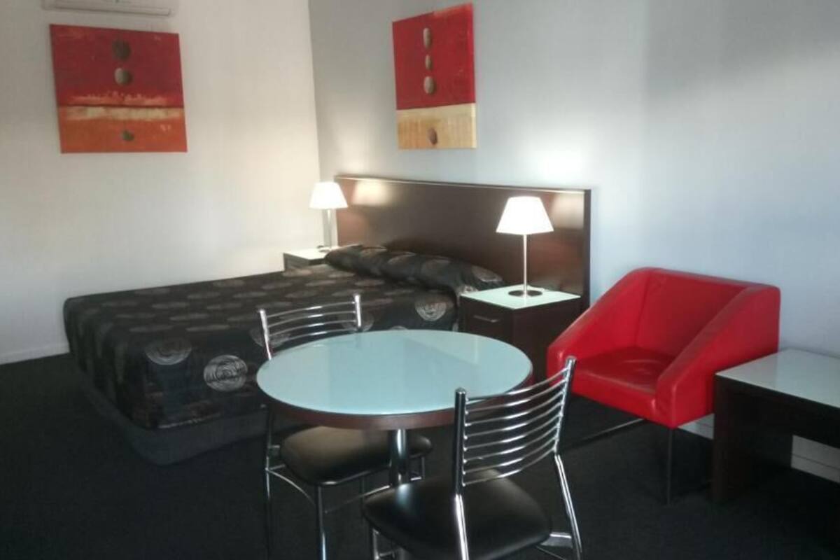 Quarto de hotel com uma cama, uma mesa redonda com duas cadeiras, uma poltrona vermelha e duas luminárias de cabeceira.