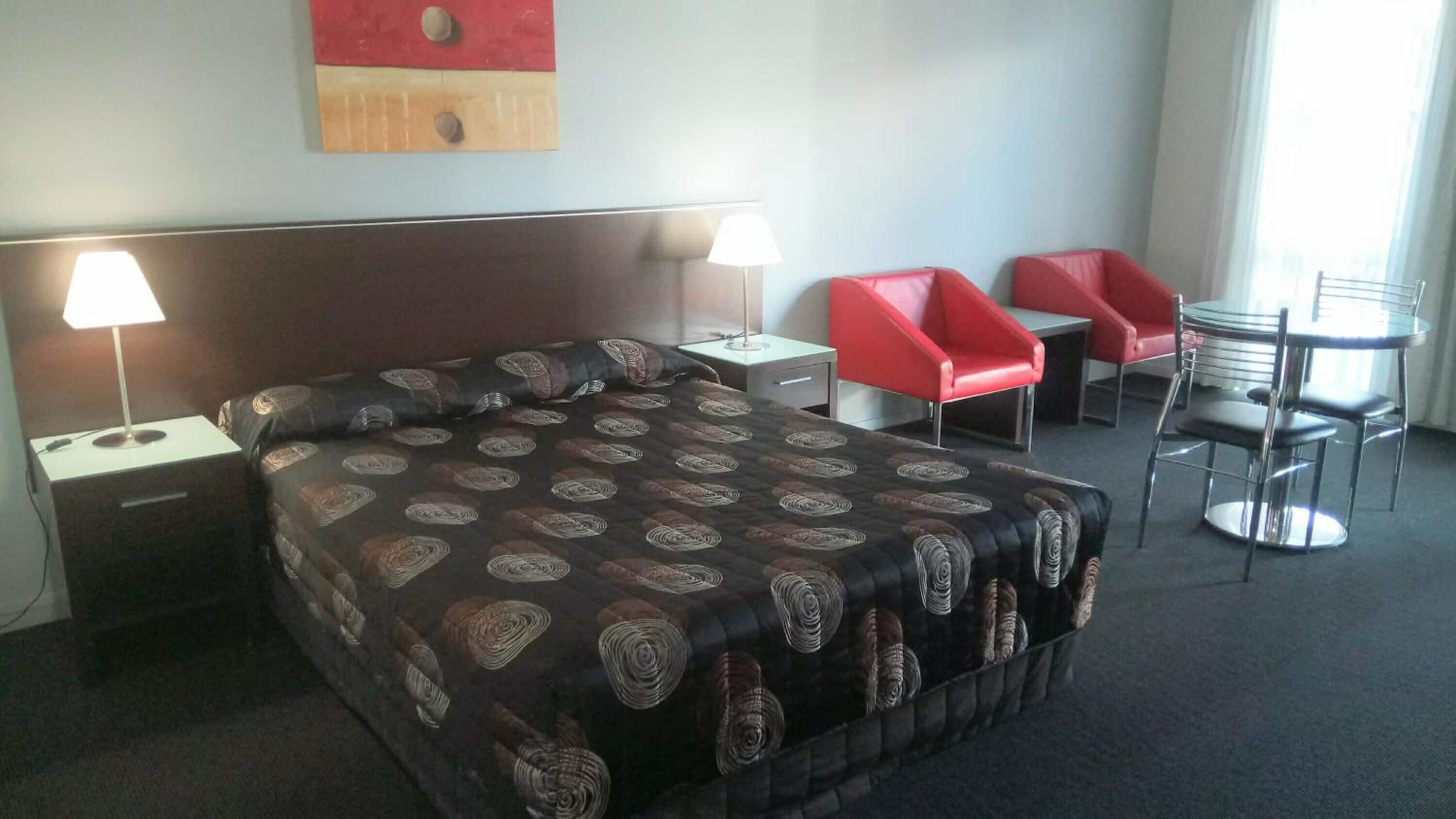 Ein Hotelzimmer mit einem großen Bett, zwei Nachttischen mit Lampen, einer Kopfteil, einem roten Sessel, einem runden Tisch mit Stühlen und einem Fenster mit Vorhängen.
