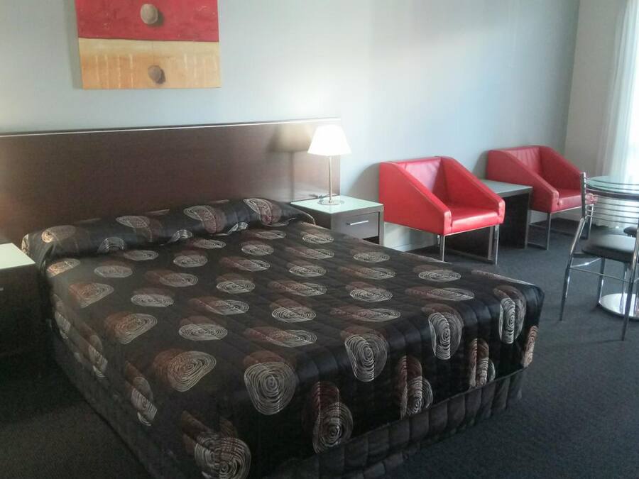 A hotel room with a large bed, two bedside tables with lamps, a headboard, a red chair, a round table with chairs, and a window with curtains.