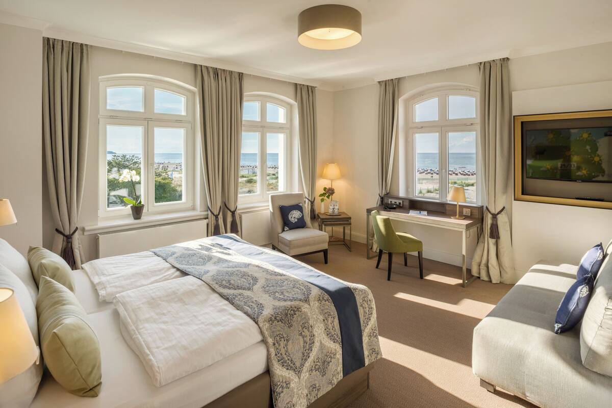A hotel room with a large bed, a desk, a chair, and a sofa. There are two windows with curtains, and a lamp on the desk.