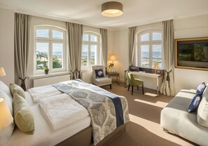A hotel room with a large bed, a desk, a chair, and a sofa. There are two windows with curtains, and a lamp on the desk. - Hotel Am Leuchtturm (Rostock)