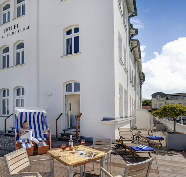 A hotel with a white facade, outdoor seating area with tables and chairs, and a person relaxing on a striped lounge chair. - Hotel Am Leuchtturm (Rostock)