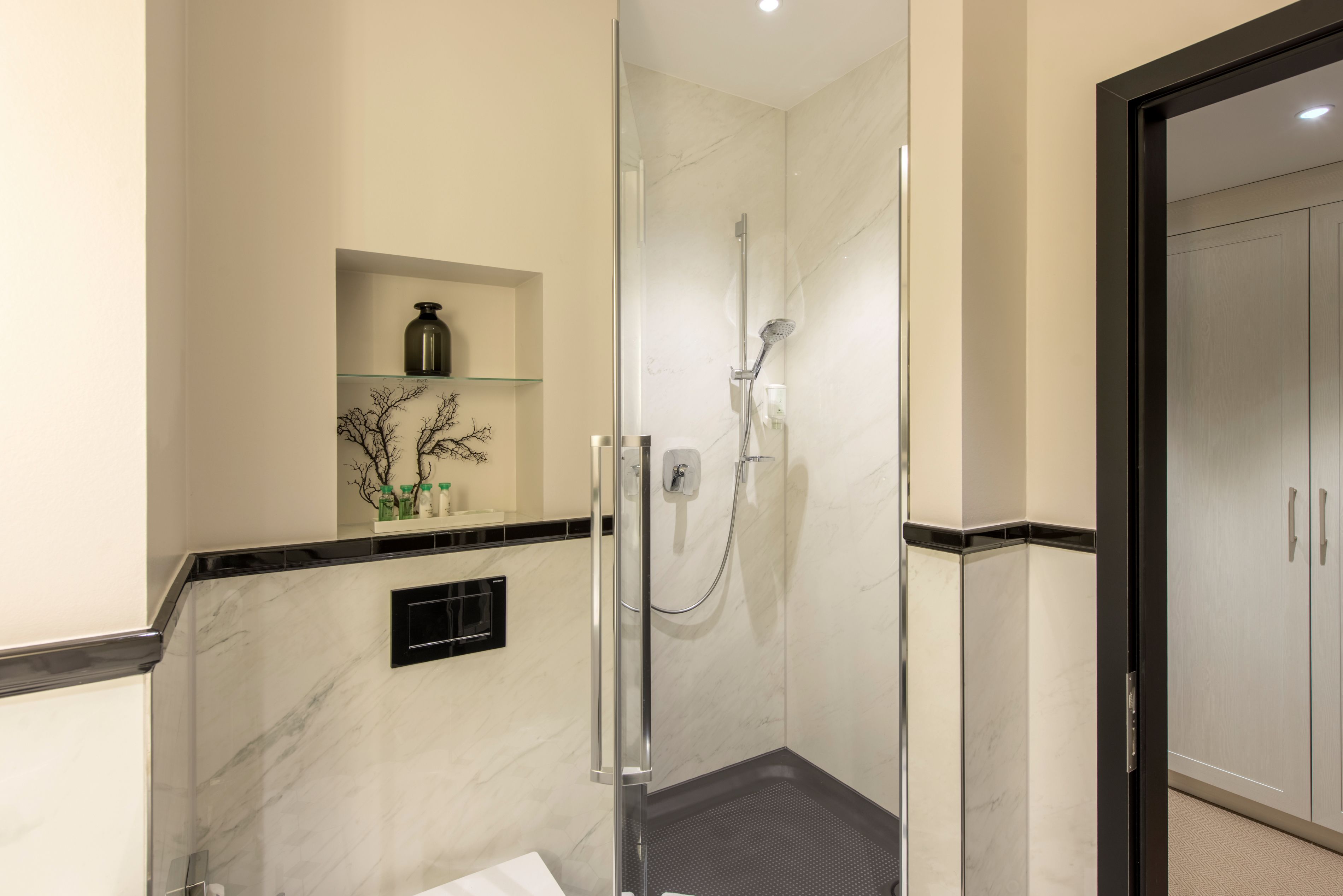 A modern bathroom with a glass-enclosed shower, marble walls, and a small shelf with toiletries.
