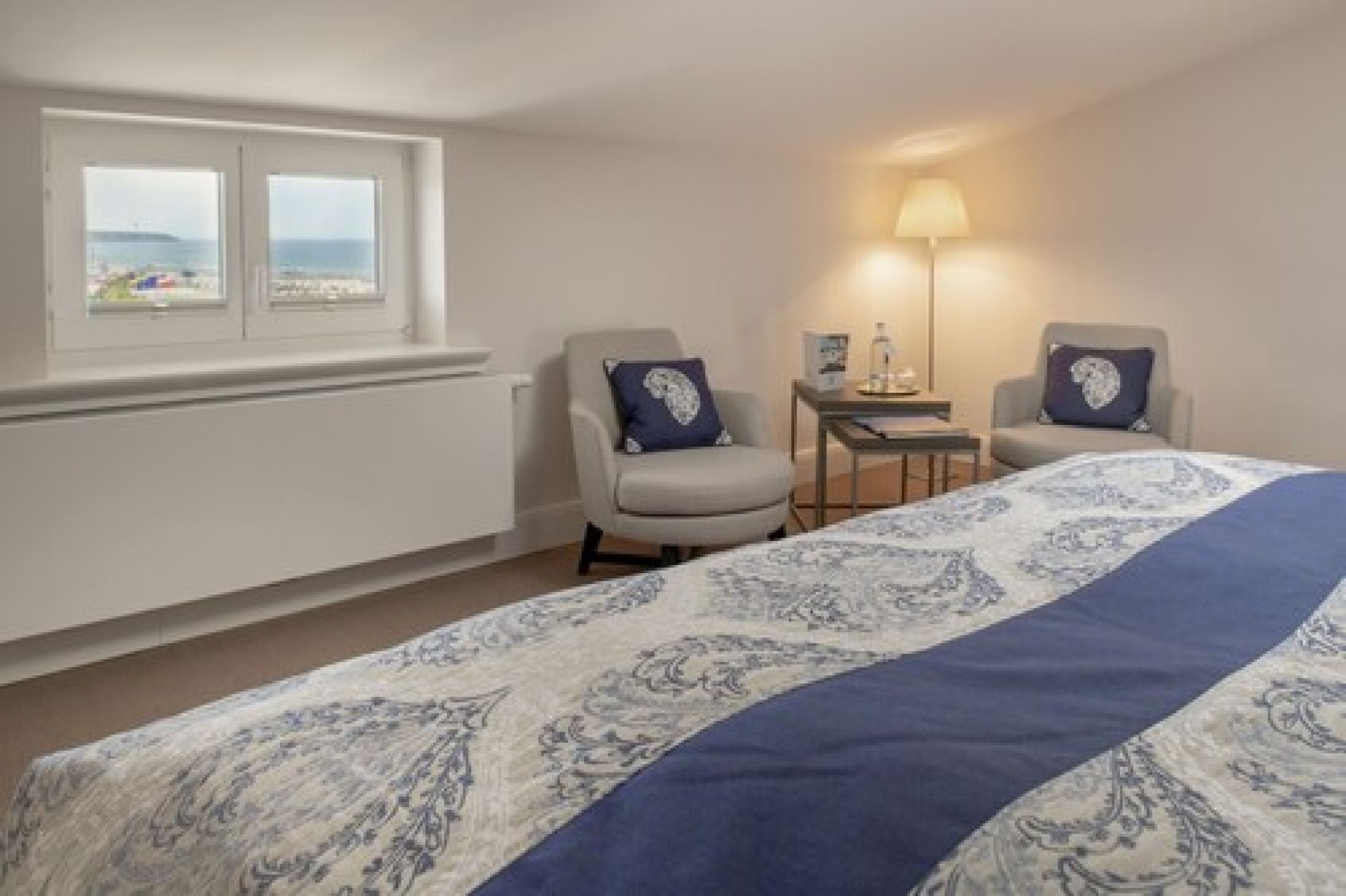 Quarto de hotel com cama, duas poltronas, uma mesinha, luminária e janela com vista para a praia.