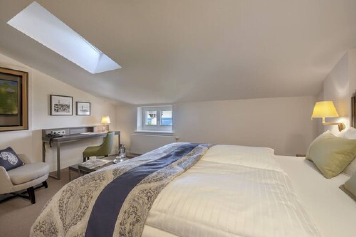 A hotel room with a bed, a desk, a chair, a lamp, and a skylight.