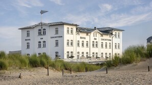 Un hotel grande y blanco con la leyenda "Hotel" y "Leuchtturm" en su fachada, ubicado en una playa arenosa con césped delantero.
