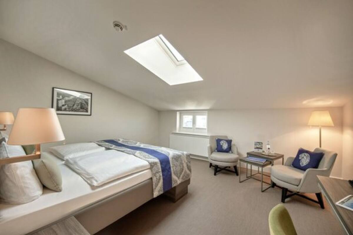 A hotel room with a bed, two chairs, a desk, a lamp, a picture on the wall, and a skylight.