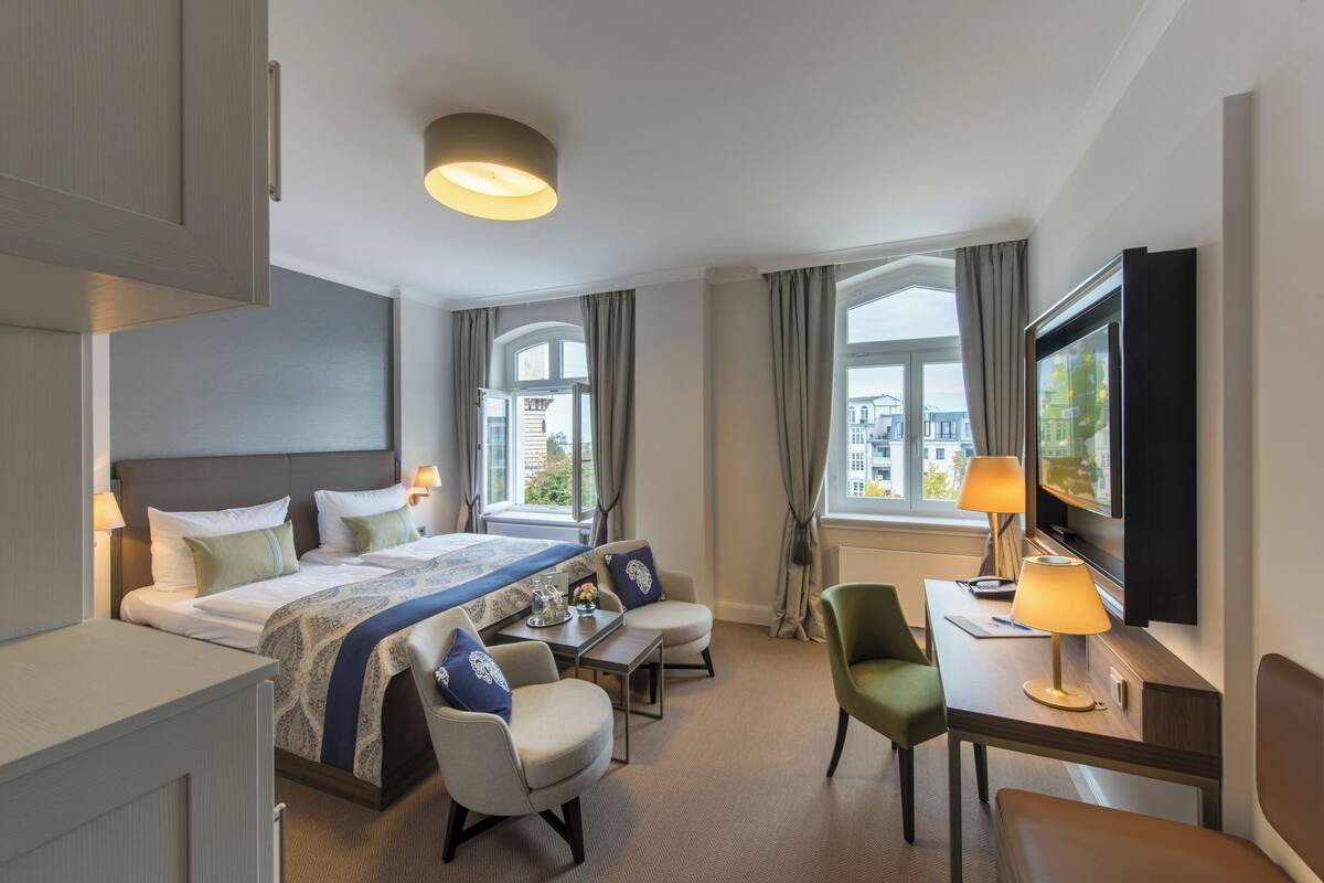 A hotel room with a bed, two chairs, a desk, a TV, and large windows.