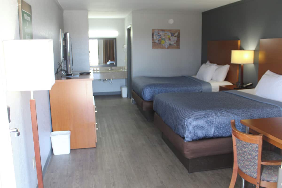 A hotel room with two beds, a desk, a chair, a small table, a mirror, a sink, and a trash bin.