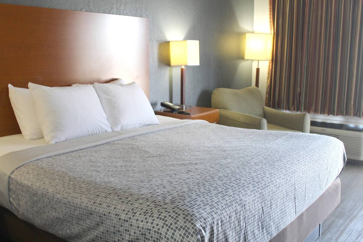 A hotel room with a large bed, a nightstand with a lamp, a chair, and a window with curtains.
